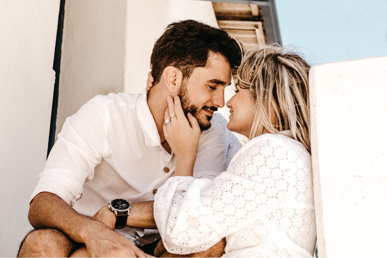 A couple sitting on steps about to share a kiss with the woman’s engagement ring in view on her hand touching his face as she smiles