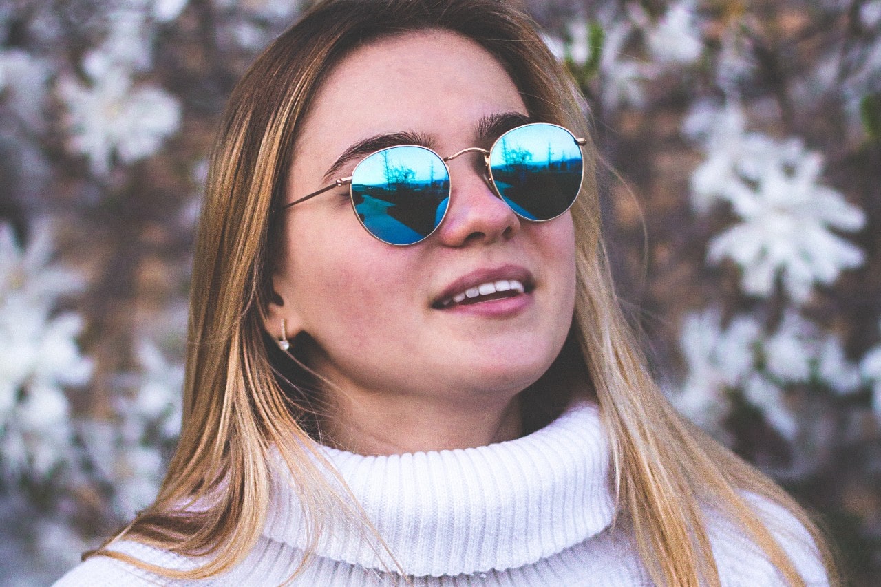 Woman with long, straight blonde hair wearing blue reflective sunglasses, a white turtleneck, and diamond huggies earrings outside