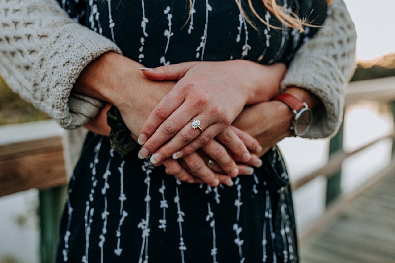 A couple standing together in a hugging embrace with the halo engagement ring the focus on her hand sitting on top of his wrapped around her